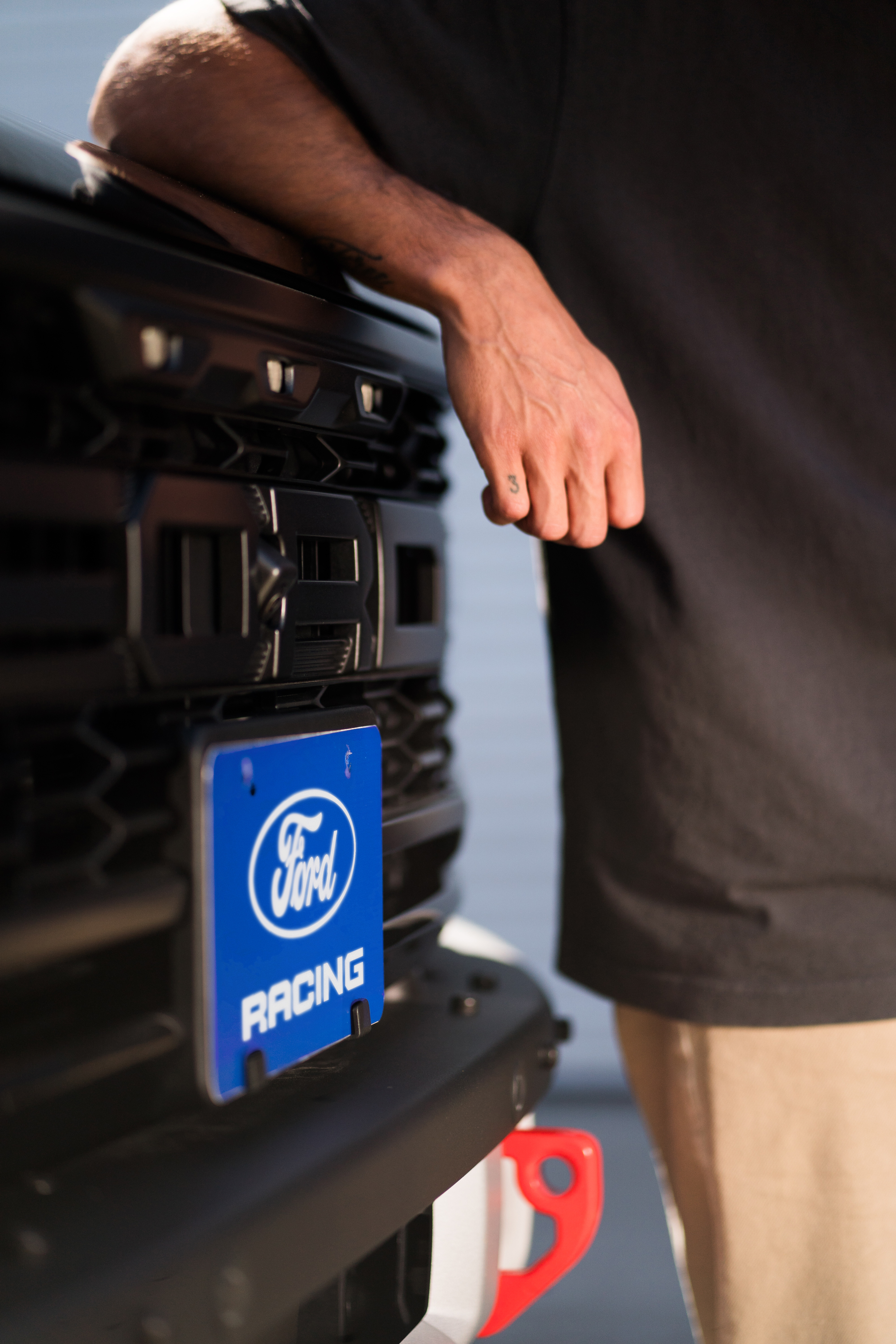 A man with a "3" tattood on his pinkie finger leans against the front grille of a truck. Only his hand, torso, and the grille with the license plate are visible. The license plate is blue and reads "Ford Racing."